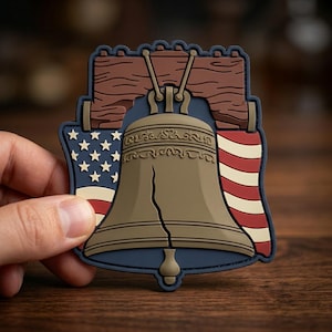 May include: A patch featuring the Liberty Bell with a crack, set against a blue background. The design includes the American flag and a brown wooden beam. The patch is held by a hand.