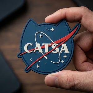 May include: A navy blue patch with a cat-shaped outline, featuring the text "CATSA" in white, and a red swoosh. The design includes white stars and a curved white line, resembling a space-themed logo.