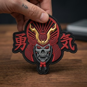 May include: A black, red, and gold morale patch featuring a stylized skull wearing a samurai helmet with golden horns. The design includes Japanese kanji characters on both sides of the helmet, set against a circular sunburst background.
