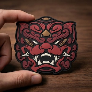 May include: A rubberized patch featuring a stylized, symmetrical tiger head design in deep red, black, and cream colors. The graphic depicts a fierce feline face with sharp fangs, intricate swirling patterns, and pointed ears on a black background.