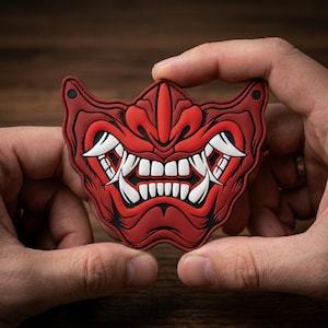 May include: A red and black rubberized patch featuring a stylized oni mask design. The mask displays prominent white fangs, a wide grin, and detailed facial contours. Small circular holes are positioned at the top corners of the patch.