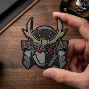 May include: A tactical morale patch featuring a stylized samurai helmet design in black, gray, and tan. The helmet has sharp, curved horns, a central hexagonal emblem, and glowing red eyes set within a dark, angular face mask structure.