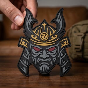 May include: A black, gray, and gold rubberized patch featuring a stylized samurai helmet design. The helmet includes a central gold emblem, dark red eyes, and intricate gray line work on a black background, shaped like a warrior mask.