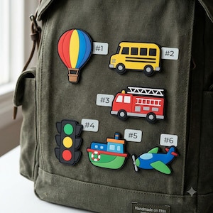 May include: An olive green canvas backpack featuring six colorful rubber patches labeled #1 through #6. The patches depict a hot air balloon, a yellow school bus, a red fire truck, a traffic light, a green and blue boat, and a blue airplane.