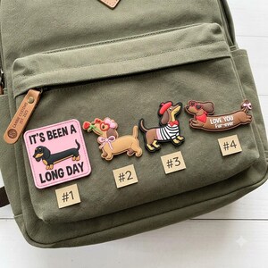 May include: An olive green canvas backpack featuring four dachshund-themed accessories. Item #1 is a pink square patch with a dachshund and the text IT'S BEEN A LONG DAY. Items #2, #3, and #4 are dachshund-shaped charms with various outfits.
