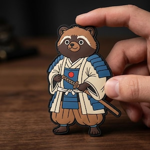 May include: A rubber patch featuring a cartoon tanuki dressed as a samurai. The tanuki has brown fur, a cream-colored kimono, blue armor plates, and a red circular emblem on the chest. It holds a katana with a brown handle and a tan scabbard.