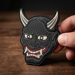 May include: A black rubberized patch shaped like a Japanese Hannya mask. The design features two curved off-white horns, wide eyes with off-white sclera, and an open mouth revealing sharp white fangs against a dark red interior and black skin.