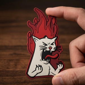 May include: A white cat patch with a red tongue and flames. The cat is screaming with its mouth open and claws out. The patch is held by a hand against a wooden background.
