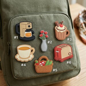 May include: Six decorative rubber charms attached to an olive green backpack. The charms include a coffee maker, a vase with red flowers, a mug with whipped cream and a cherry, a teacup on a saucer, a picnic basket, and a red toaster, labeled #1-#6.