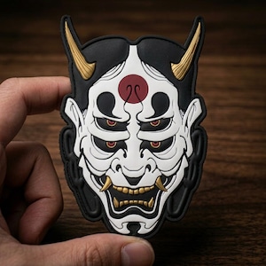 May include: A multi-eyed Hannya mask patch featuring a white face with black accents, a red circular mark on the forehead, and gold-colored horns and teeth. The design is rendered in a raised, molded style with a black border and detailed lines.