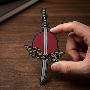 May include: A rubberized patch featuring a vertical katana sword with a black and gold handle, positioned over a solid red circle and stylized black clouds with gold outlines. The design is flat and graphic, showcasing a sharp blade and hilt.