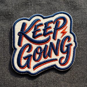 May include: Embroidered patch with the words "KEEP GOING" in a stylized font. The text is in blue with orange accents and a white background, outlined in blue. A lightning bolt graphic is above the letter "P".