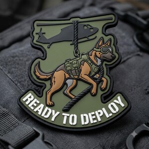 May include: An olive green tactical morale patch featuring a black silhouette of a helicopter, a German Shepherd dog wearing a tactical vest and goggles, and a rope. The text READY TO DEPLOY is printed in white at the bottom of the patch.