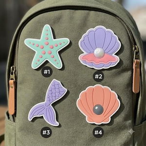 May include: Four decorative patches attached to an olive green backpack. Item #1 is a mint green starfish with pink dots. Item #2 is a purple seashell with a white pearl. Item #3 is a purple mermaid tail. Item #4 is a peach seashell with a gray pearl.