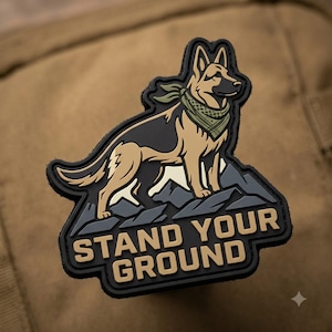 May include: A black, tan, and olive green rubber patch featuring a German Shepherd dog wearing a bandana, standing atop stylized gray mountain peaks. The text STAND YOUR GROUND is embossed in tan lettering at the bottom of the patch.