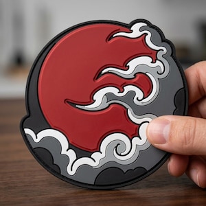 May include: A circular rubber patch featuring a bold red sun design accented with stylized white and grey clouds. The graphic is set against a black background with a raised, textured surface, creating a layered appearance with distinct color sections.