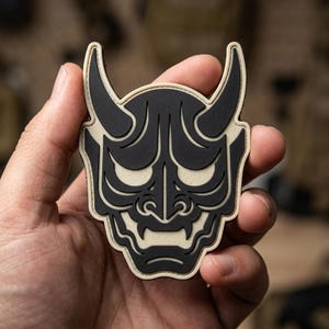May include: A morale patch featuring a stylized Hannya mask design. The patch is constructed with a matte black raised graphic layer set against a flat, light beige background, showcasing sharp horns, a wide nose, and a fierce facial expression.
