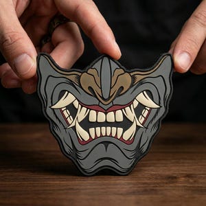 May include: A gray, rubberized patch shaped like a samurai-style facial mask. The design features prominent, cream-colored fangs, a set of teeth, and dark red accents around the mouth area, all set against a dark gray, contoured background.