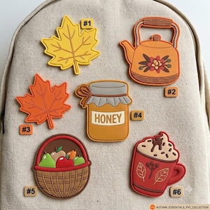 May include: Six decorative PVC charms featuring autumn themes: a yellow maple leaf #1, a tea kettle #2, an orange maple leaf #3, a honey jar labeled HONEY #4, a basket of fruit #5, and a mug with whipped cream #6, all arranged on a beige fabric surface.