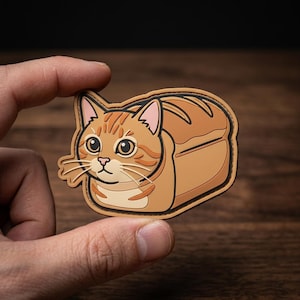 May include: A whimsical patch featuring a cat designed to look like a loaf of bread. The cat has orange and brown stripes, with a tan bread-like exterior. The patch is held by a hand.