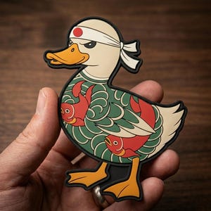 May include: A rubber patch shaped like a duck wearing a white headband with a red circle. The duck has a cream-colored head, orange beak and feet, and a green body featuring a pattern of red fish and swirling lines with a black outline border.