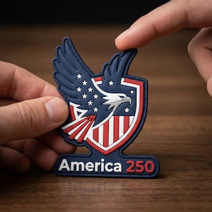 May include: A patriotic patch featuring a blue eagle with white stars and a red and white striped shield. The patch has the text "America 250" in red. The patch is held between two fingers.