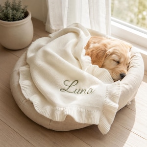 Peut inclure: Une couverture tricotée de couleur crème avec un bord à volants et le nom Luna brodé en fil vert, drapée sur un chiot doré endormi reposant dans un panier pour animal de compagnie rond en tissu beige. La couverture présente un tricot texturé.