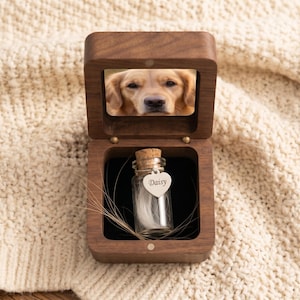 May include: A wooden pet memorial box with a photo of a golden retriever. Inside, a small glass vial holds fur and a heart-shaped tag engraved with "Daisy." The box is open, revealing a black interior and a cork-topped vial.