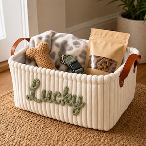 Puede incluir: Una cesta de almacenamiento rectangular de pana color crema con asas de cuero marrón y la palabra Lucky bordada en hilo verde. Dentro hay un juguete de hueso de ganchillo color canela, una manta gris y blanca con manchas, un collar verde y premios.