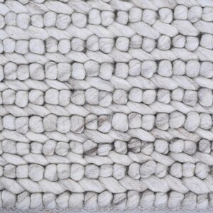 May include: Close-up of a textured, off-white rug. The rug features a thick, braided design with horizontal rows of rounded, interwoven strands. Subtle gray and brown variations add depth to the neutral color palette. The rug's construction suggests a durable, cozy floor covering.