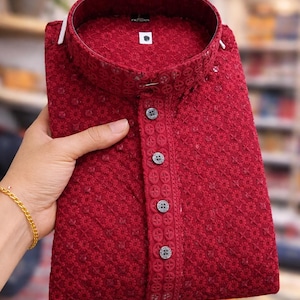 Puede incluir: Una camisa kurta roja oscura doblada con intrincados bordados florales. La camisa presenta un cuello mandarín y una tapeta abotonada con botones decorativos. La tela tiene una apariencia texturizada y el estilo general es tradicional.