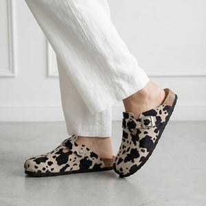 Puede incluir: Zuecos con estampado de vaca y correas de hebilla ajustables. Los zapatos sin cordones presentan un estampado de vaca en blanco y negro sobre un fondo beige, con suela de corcho y suela de goma negra.
