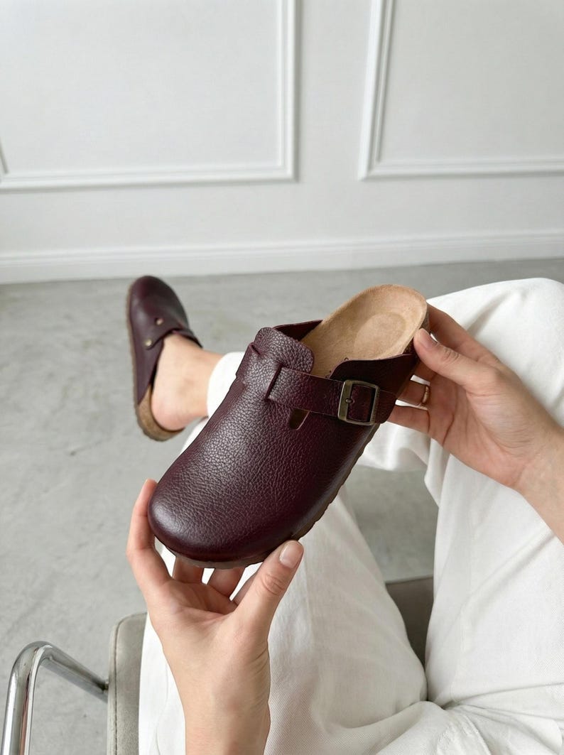 May include: A pair of dark burgundy clogs with a textured leather upper, a rounded toe, and an adjustable strap featuring a bronze-toned metal buckle. The interior footbed is light tan, and the sole is constructed from a natural cork material.