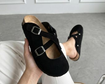 May include: A pair of black suede clogs featuring a rounded toe, a cork-style footbed, and two adjustable crisscross straps across the top. Each strap is secured with a silver-toned metal buckle, providing a functional closure for the footwear.