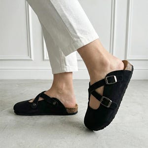 Op de afbeelding: Zwarte suède instappers met twee verstelbare gespriemen. De sandalen hebben een kurken voetbed. Het schoeisel is ontworpen voor comfort en casual wear.