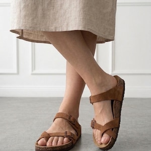 May include: Brown suede sandals featuring a toe loop design, two adjustable straps with metal buckles, and a contoured cork footbed. The footwear has a dark brown textured outsole and a slip-on construction with an open toe and open heel.