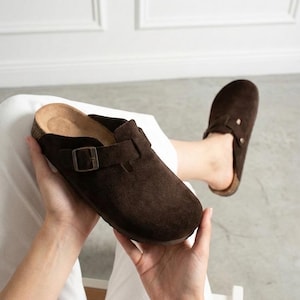 May include: A pair of dark brown suede clogs featuring a rounded toe, a cork-textured footbed, and an adjustable strap across the top with a metallic buckle. The footwear has a slip-on design with a closed front and an open back construction.