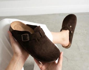 May include: A pair of dark brown suede clogs featuring a rounded toe, a cork-textured footbed, and an adjustable strap across the top with a metallic buckle. The footwear has a slip-on design with a closed front and an open back construction.