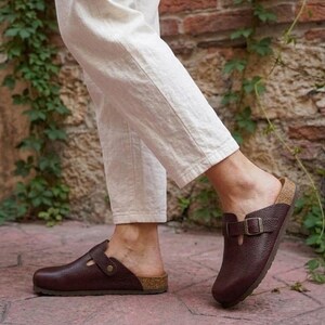 May include: A pair of dark brown leather clogs featuring a textured finish, a rounded toe, and an adjustable strap across the top with a metal buckle. The footwear has a cork-style midsole and a flat sole, worn with off-white linen trousers.