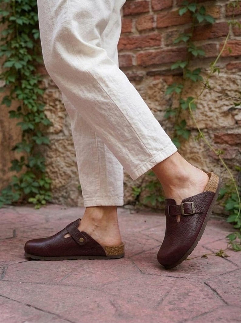 May include: A pair of dark brown leather clogs featuring a textured finish, a rounded toe, and an adjustable strap across the top with a metal buckle. The footwear has a cork-style midsole and a flat sole, worn with off-white linen trousers.