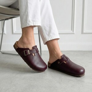May include: A pair of dark burgundy leather clogs featuring a textured finish, a rounded toe, and a single adjustable strap across the top with a metallic buckle. The footwear has a visible cork-style midsole and a dark brown outsole.