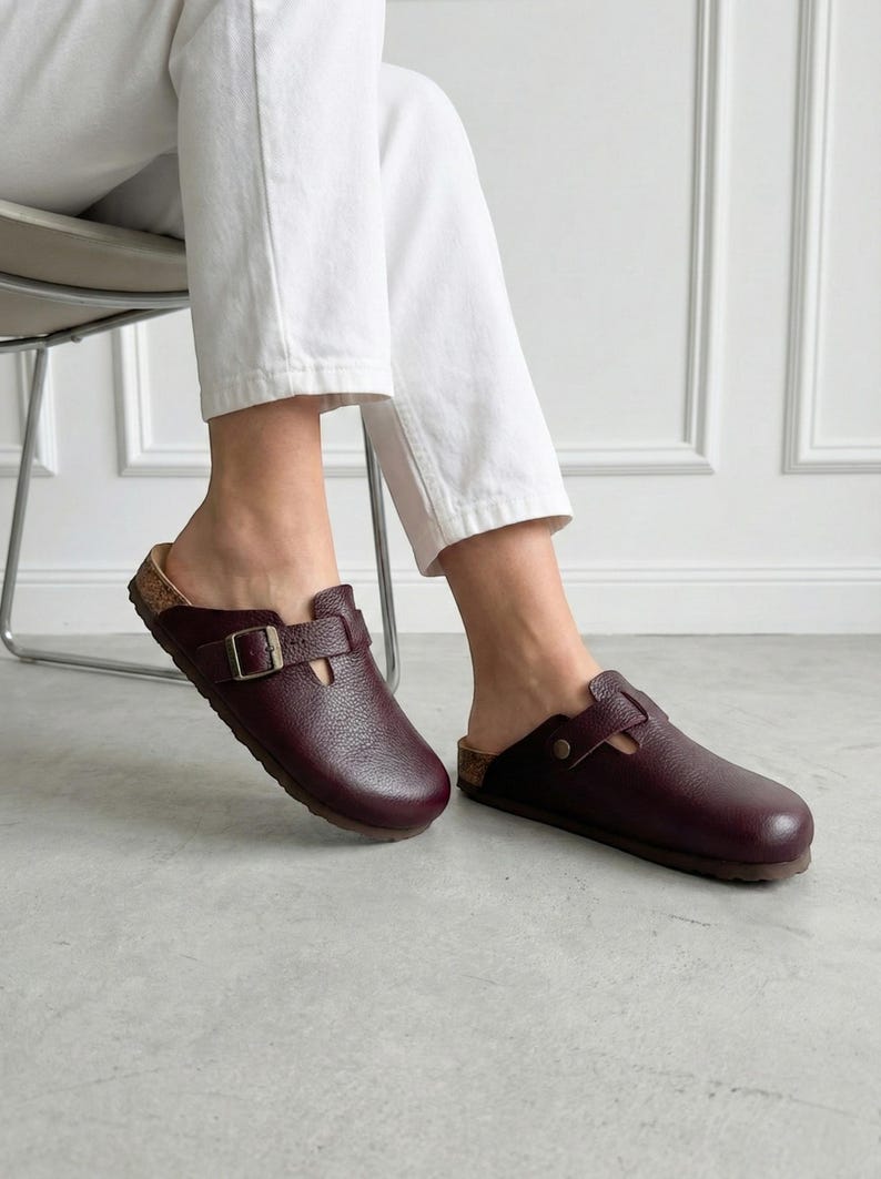May include: A pair of dark burgundy leather clogs featuring a textured finish, a rounded toe, and a single adjustable strap across the top with a metallic buckle. The footwear has a visible cork-style midsole and a dark brown outsole.