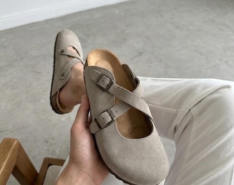 May include: A pair of taupe suede clogs featuring a closed round toe, a cork footbed, and two adjustable straps with bronze-toned metal buckles that cross over the top of the foot. The footwear has a sturdy sole and a soft, matte exterior finish.