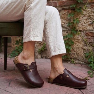 May include: A pair of dark brown leather clogs featuring a textured grain finish, a rounded toe, and a single adjustable strap across the instep secured with a metallic buckle. The footwear has a cork-style midsole and a flat, dark brown outsole.