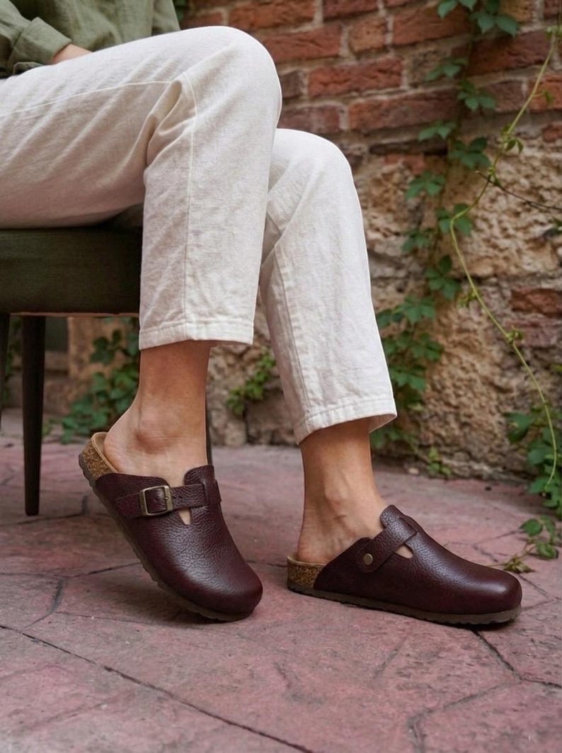 May include: A pair of dark brown leather clogs featuring a textured grain finish, a rounded toe, and a single adjustable strap across the instep secured with a metallic buckle. The footwear has a cork-style midsole and a flat, dark brown outsole.