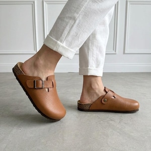 Può includere: Zoccoli in pelle marrone con fibbia. Gli zoccoli hanno un plantare in sughero e un design a punta chiusa. Le scarpe sono di colore marrone caldo e sembrano essere realizzate in pelle strutturata. Gli zoccoli sono indossati con pantaloni bianchi.