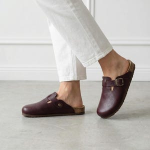 May include: A pair of dark burgundy leather clogs featuring a rounded toe, a textured grain finish, and an adjustable strap across the top secured with a metallic buckle. The footwear has a cork-style midsole and a flat, dark brown outsole.