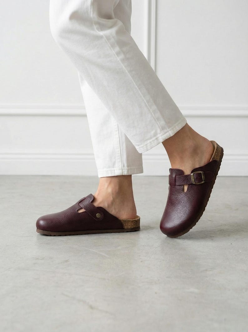 May include: A pair of dark burgundy leather clogs featuring a rounded toe, a textured grain finish, and an adjustable strap across the top secured with a metallic buckle. The footwear has a cork-style midsole and a flat, dark brown outsole.