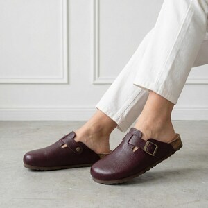 May include: A pair of dark burgundy leather clogs featuring a textured finish, a rounded toe, and a single adjustable strap across the top secured with a metallic buckle. The footwear has a cork-style footbed and a dark brown sole.