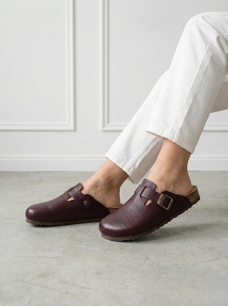 May include: A pair of dark burgundy leather clogs featuring a textured finish, a rounded toe, and a single adjustable strap across the top secured with a metallic buckle. The footwear has a cork-style footbed and a dark brown sole.